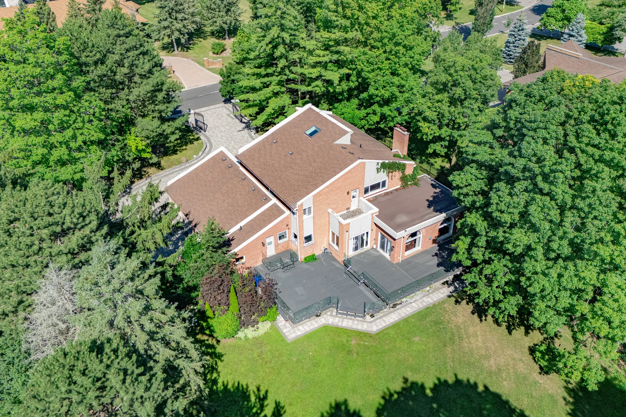 The back of the house from above: brown roofs with a skylight and a chimney, the large grey deck with its glass railing, the lawn and tall trees
