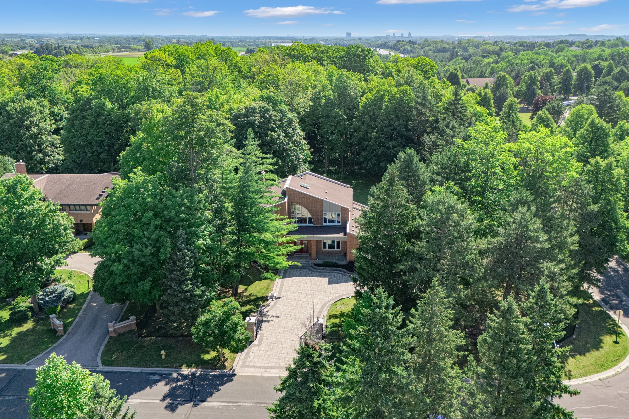 The house from above the street: brick walls and brown roofs among tall pines and maples, the paved drive and the gate at the curb, the street curving past