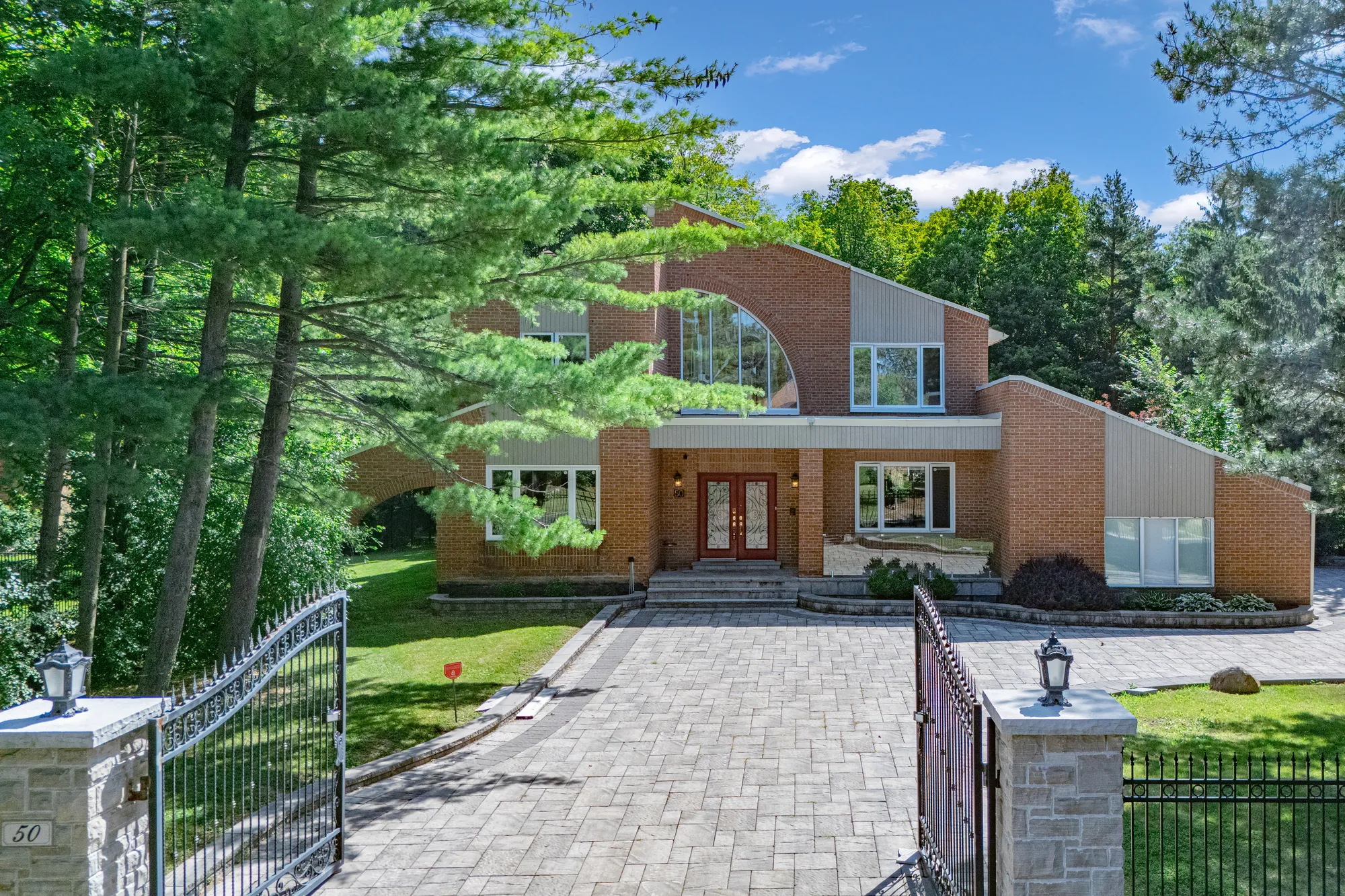 From above the street: the stone gate pillars with lanterns and the house number 50, the iron gates open onto the paved drive, the front of the house beyond under tall pines