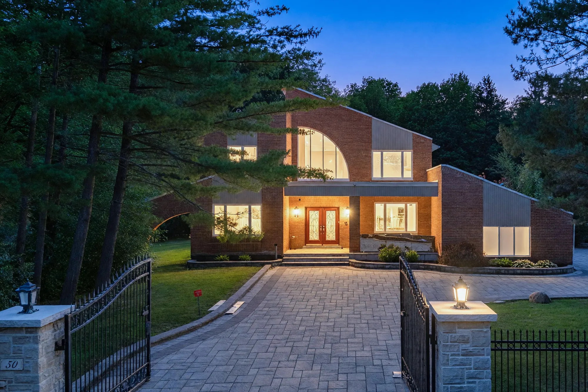 The same view at dusk: the lantern on the right gate pillar lit, the windows and the covered entry of the house lit beyond the drive
