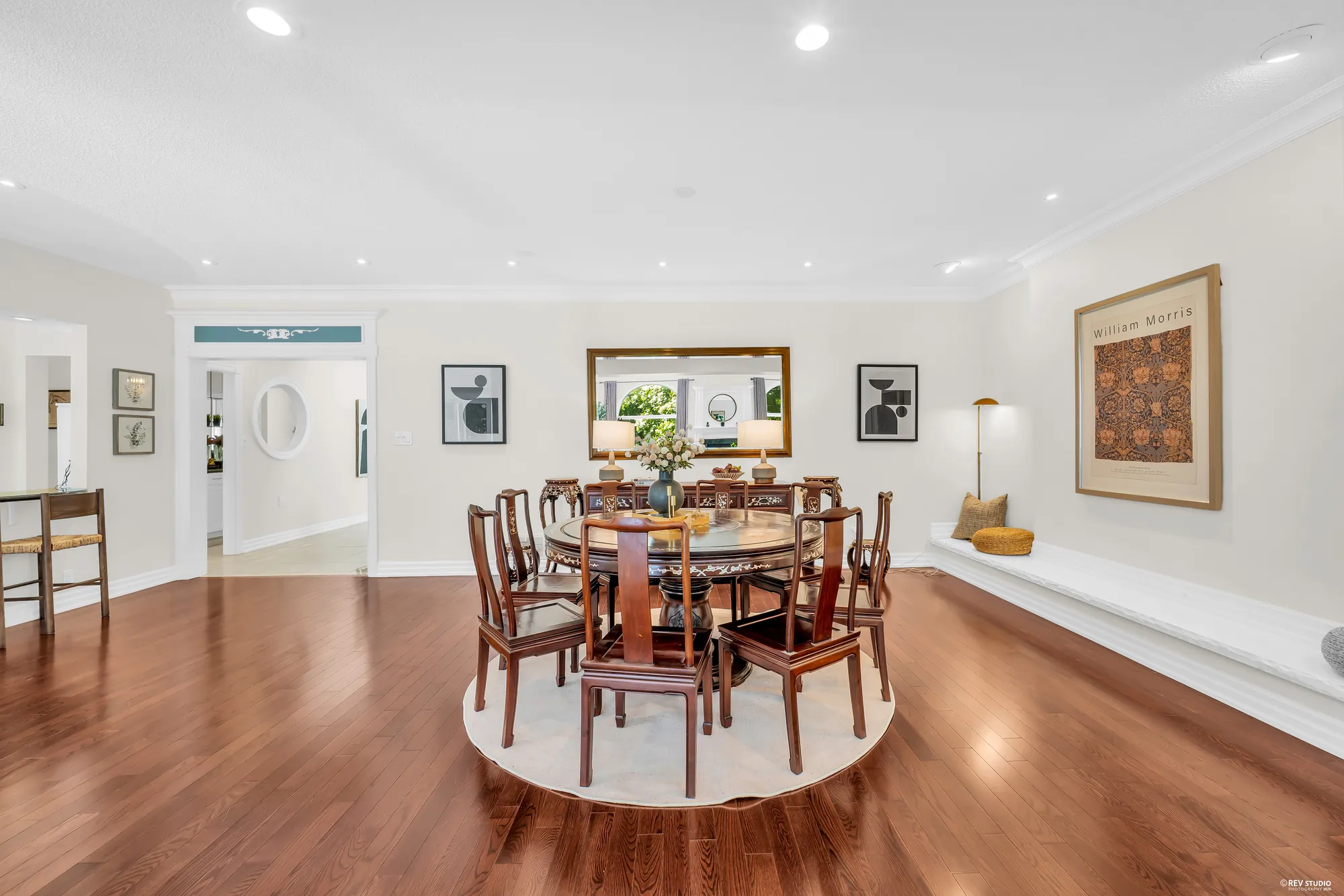 The dining area at the back: a round wood table with carved chairs on a round rug, framed prints, a mirror over a sideboard, a built-in bench along the wall