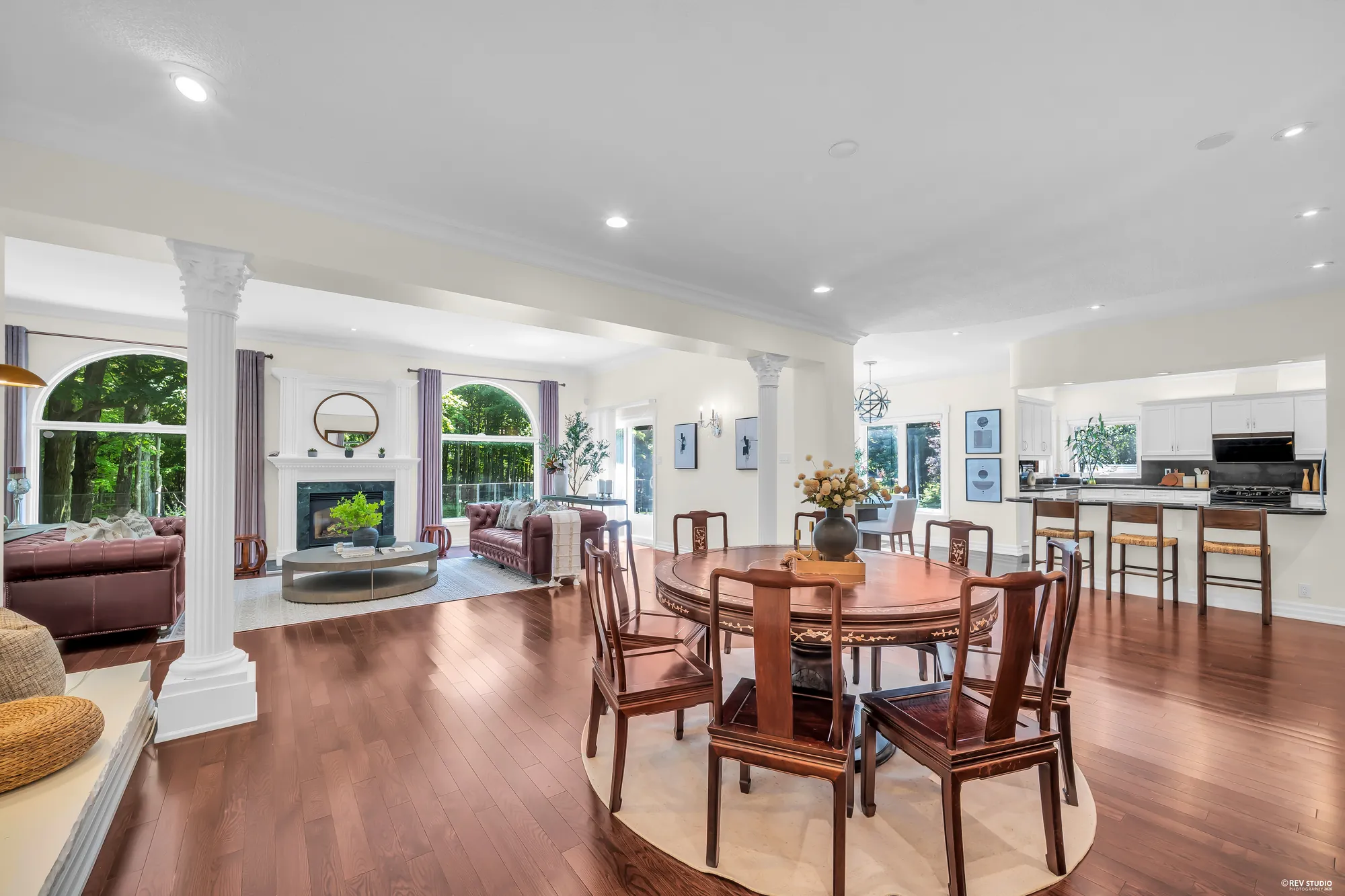 The round dining table between white columns, the family room and its fireplace at the left, the kitchen&rsquo;s breakfast bar at the right