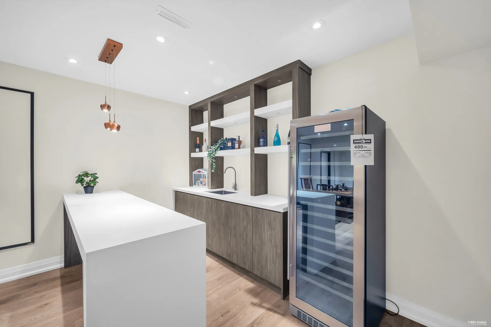 The wet bar: a white island, a counter with a sink, open shelving in dark wood, a wine fridge, copper pendant lights