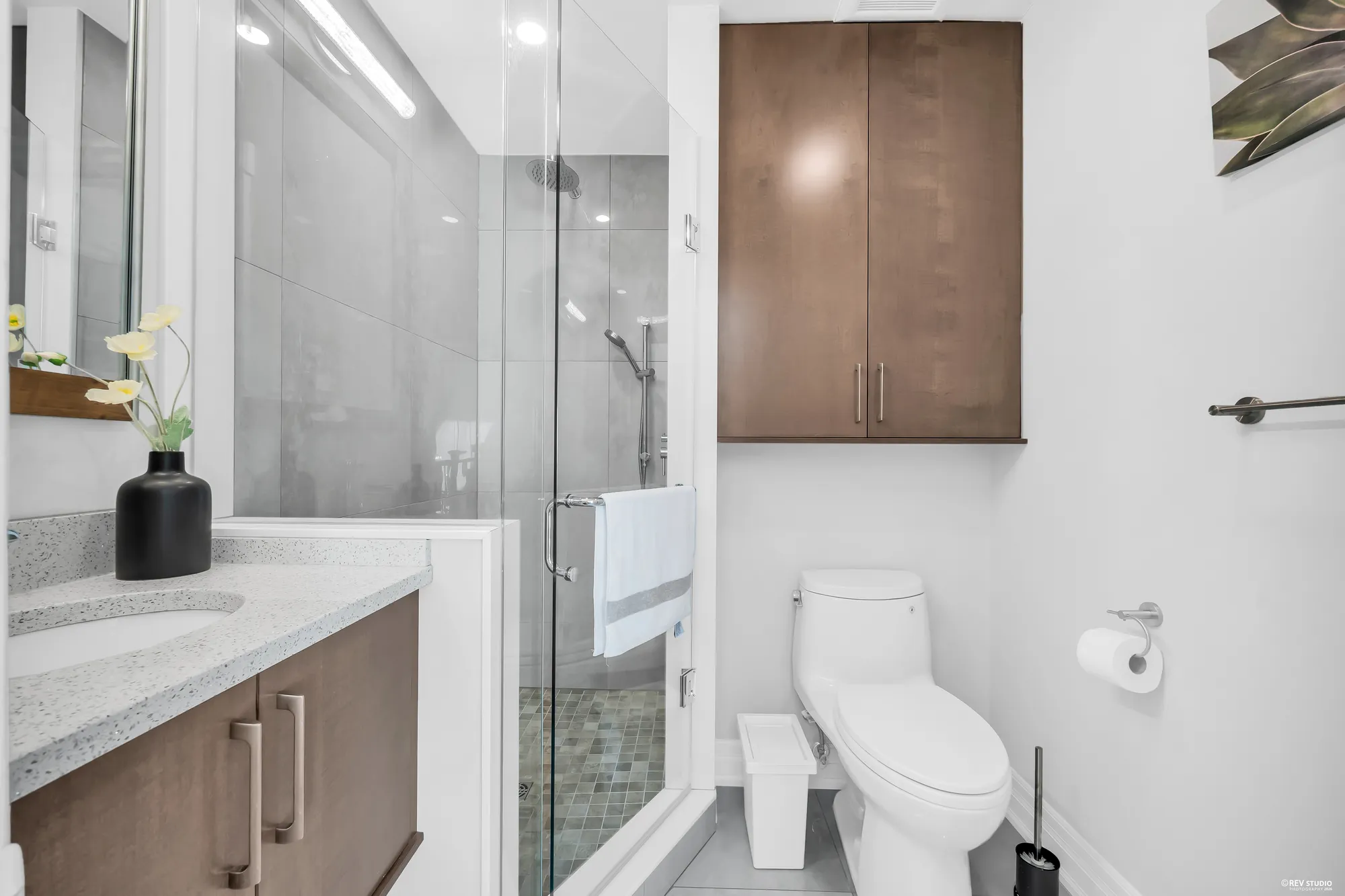 A bathroom: a glass shower in large grey tile, a vanity with a speckled quartz top, a toilet, a wood cabinet above