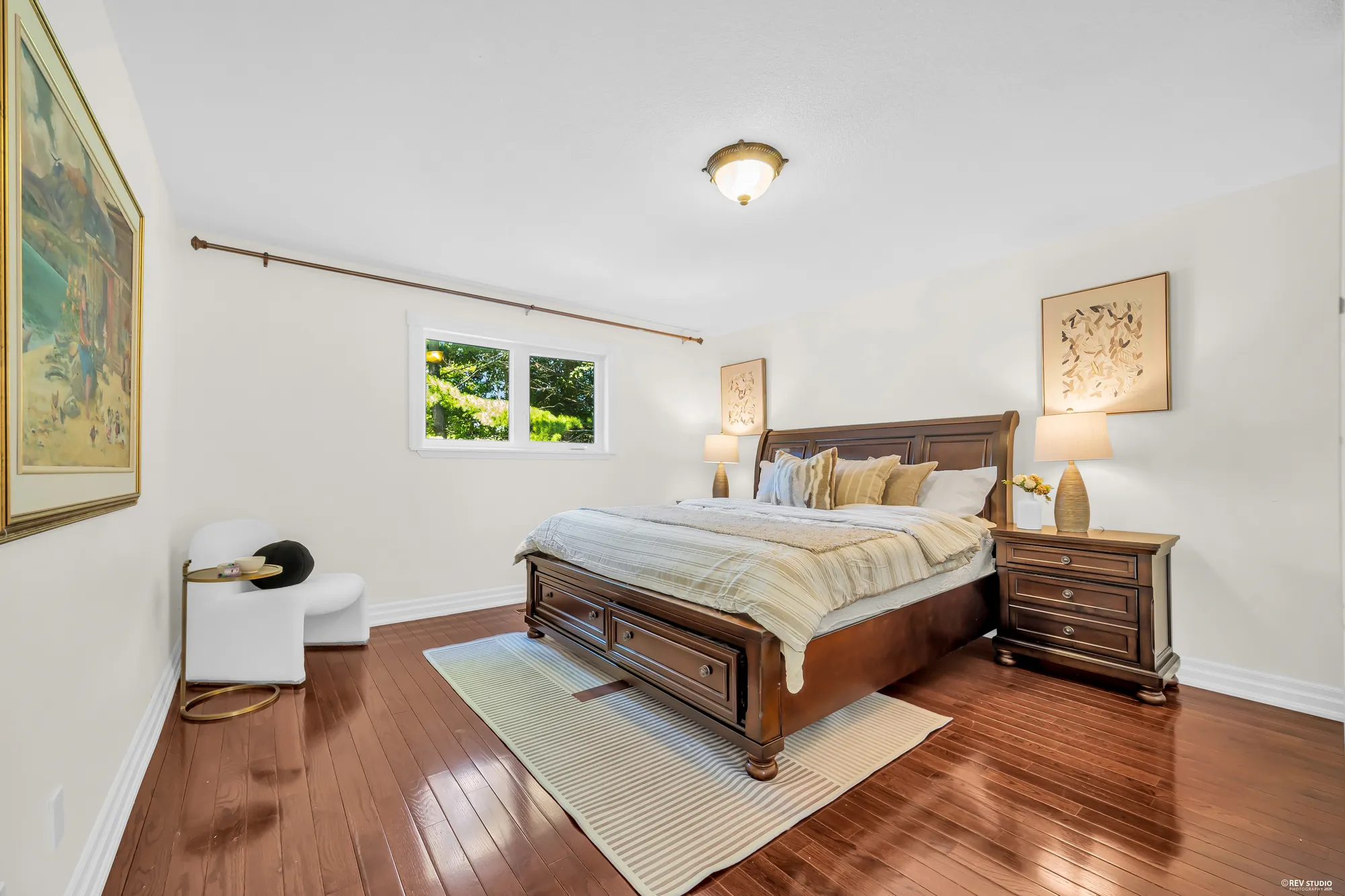 A bedroom: a wood bed with storage drawers, night tables with lamps, a window, a large framed painting on the wall, a white chair