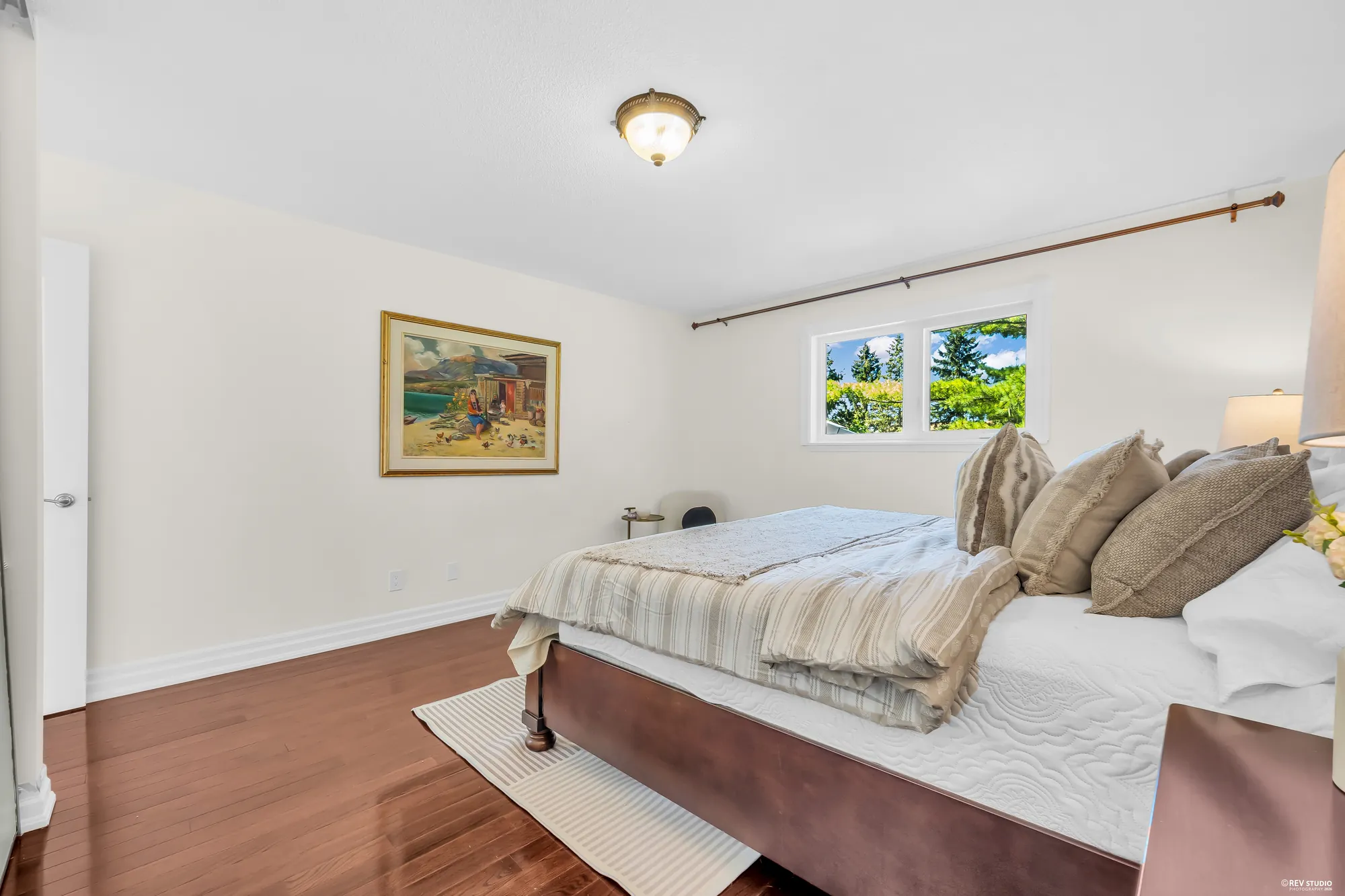 The same bedroom: the bed with striped bedding, a framed painting of a shore, a window onto the trees