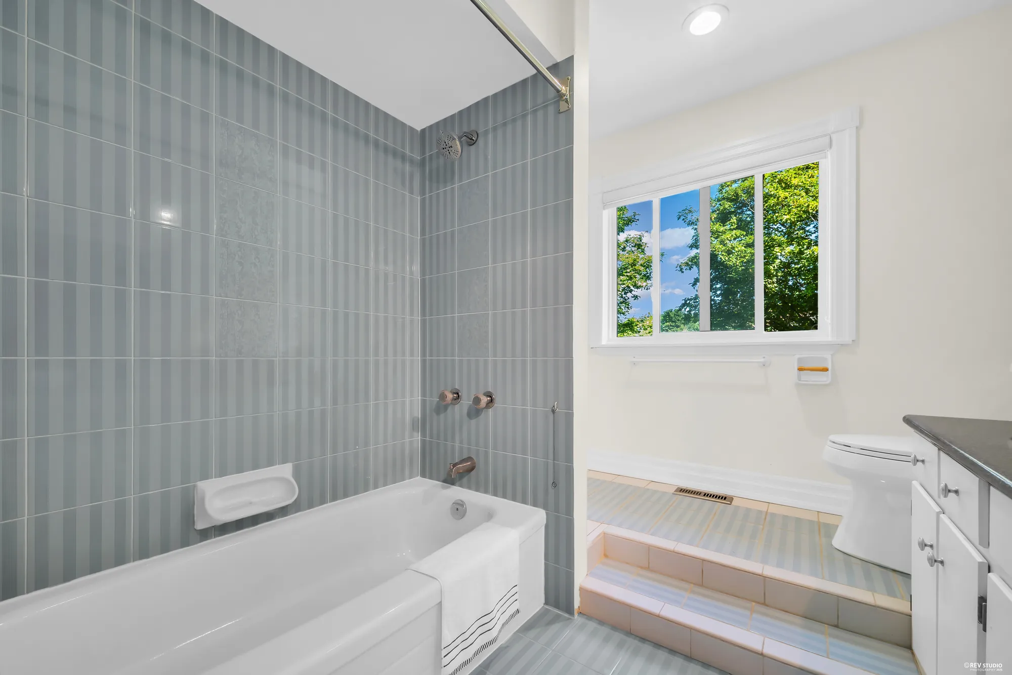 A bathroom: a tub with a shower in blue-grey tile, a window, a toilet