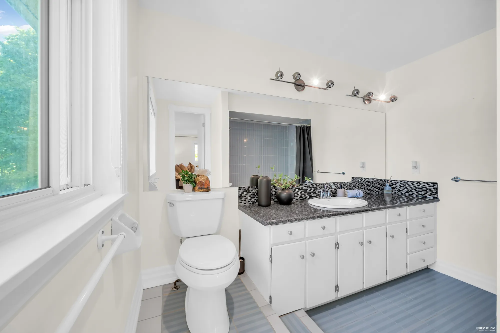 The same bathroom: a white vanity with a granite top and a mosaic backsplash, a toilet, a window and a grab bar