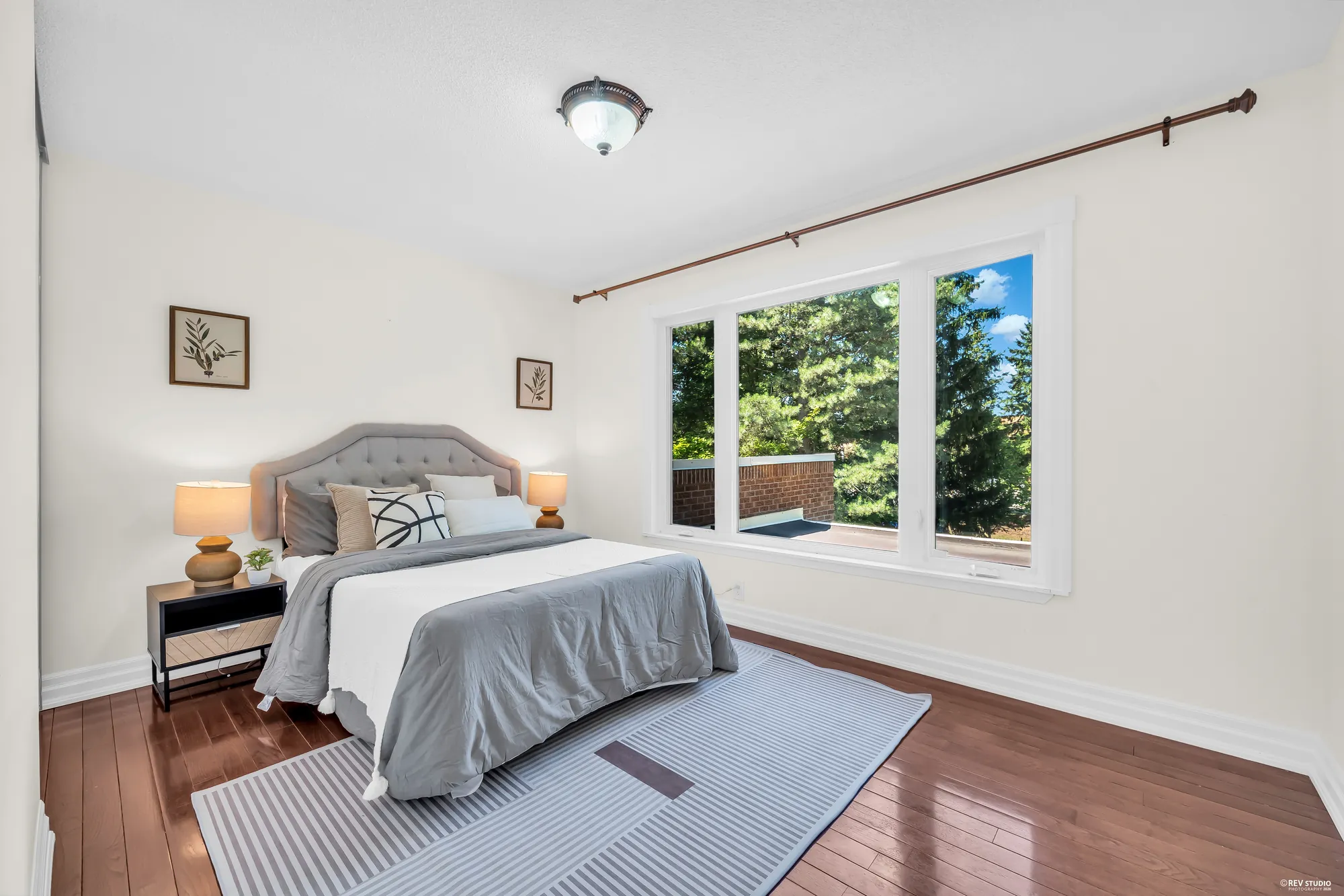 A bedroom: a grey upholstered headboard, night tables with lamps, a wide three-part window onto the trees and a flat roof below