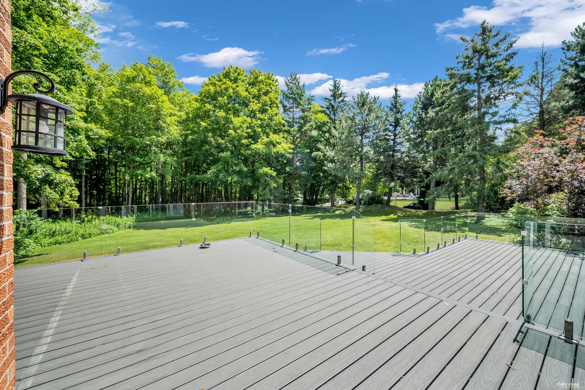 The deck from beside the house: grey composite boards, a glass railing on posts, a lantern on the brick wall, the lawn and a line of tall trees
