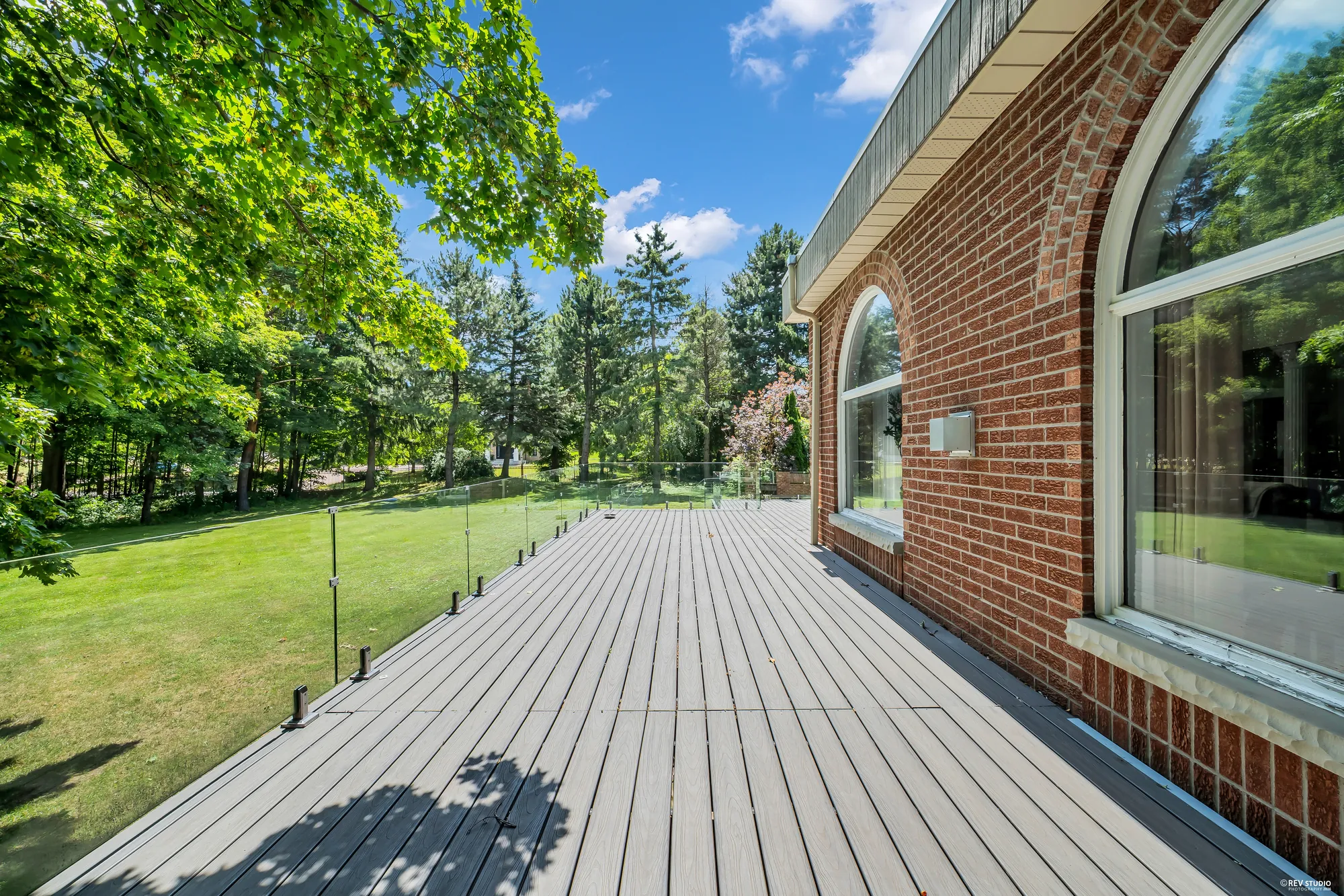 The deck along the back of the house: grey composite boards, a glass railing, a brick wall with tall arched windows, the lawn and trees