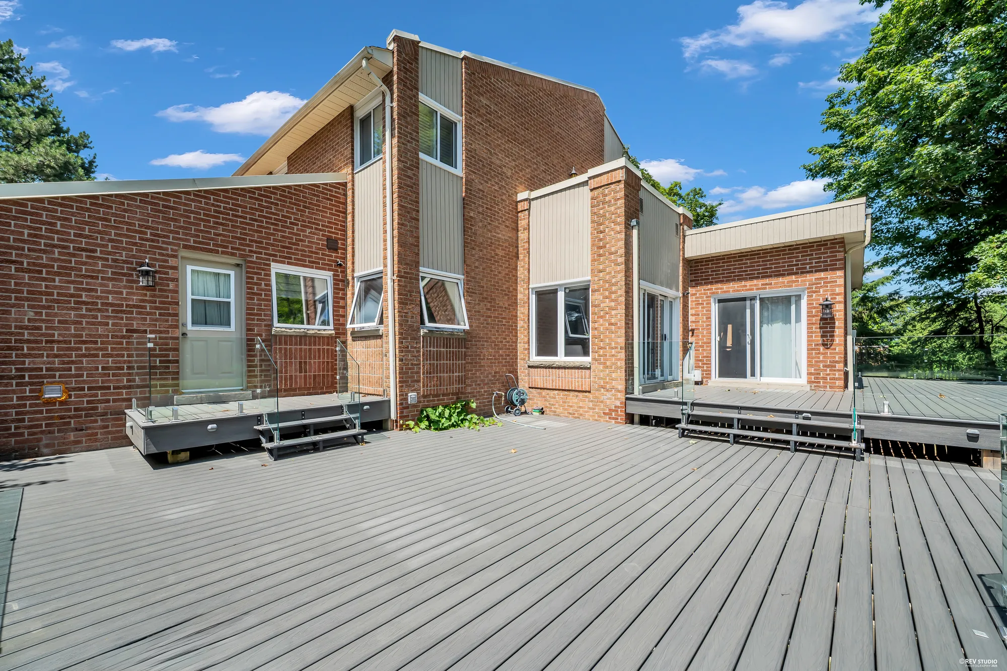 The back of the house from the deck: red brick and grey siding on two storeys, a door and windows, steps up to a raised part of the deck