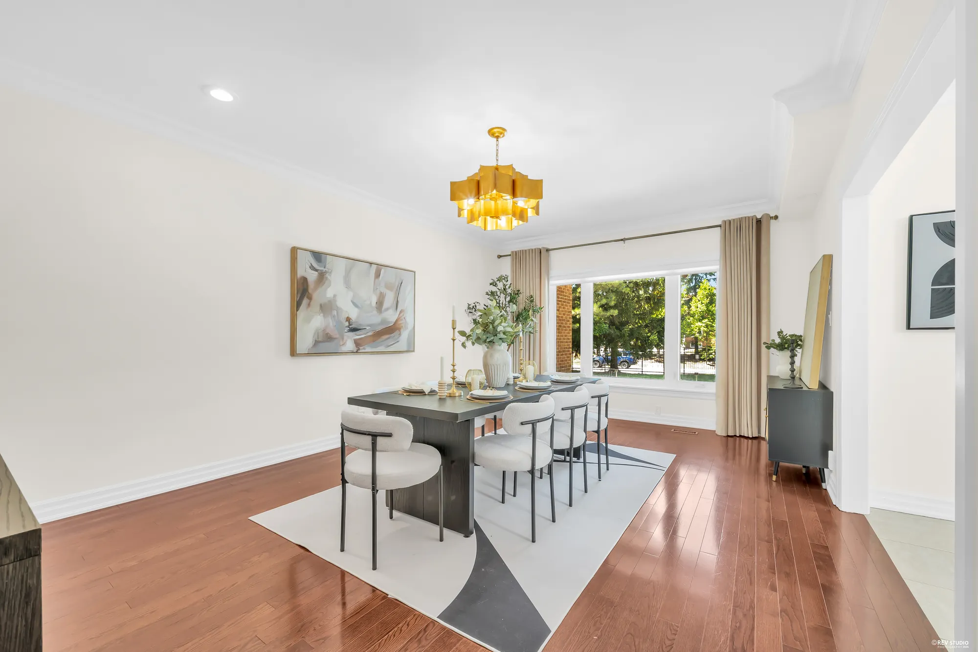 The front dining room: a dark table with cream chairs under a gold pendant light, an abstract painting, a window onto the front lawn