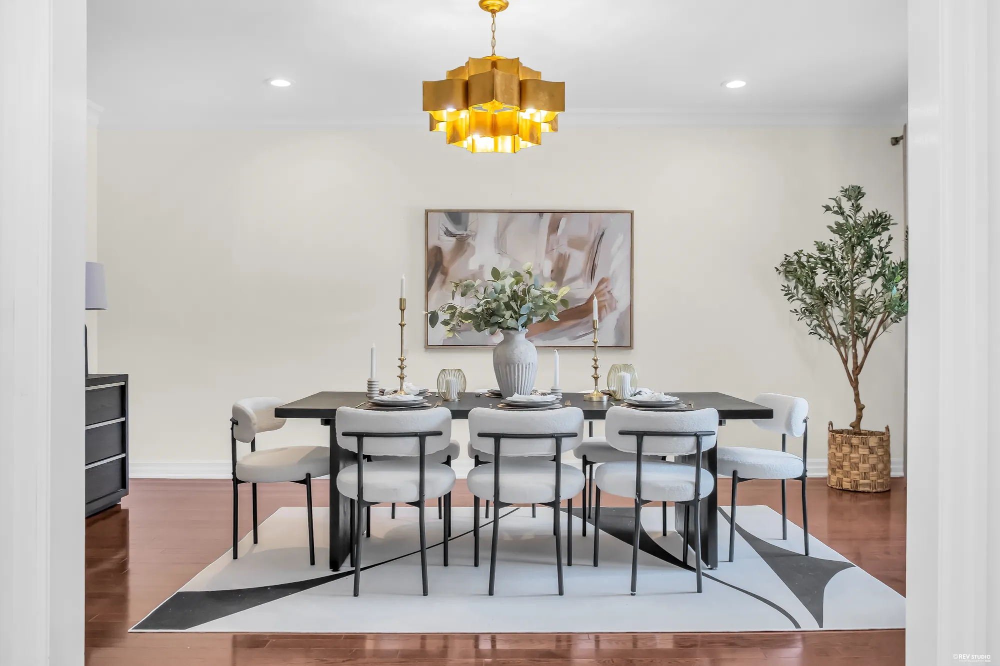 The table set with plates and candlesticks, cream chairs, a rug in black and grey, a large print and an olive tree