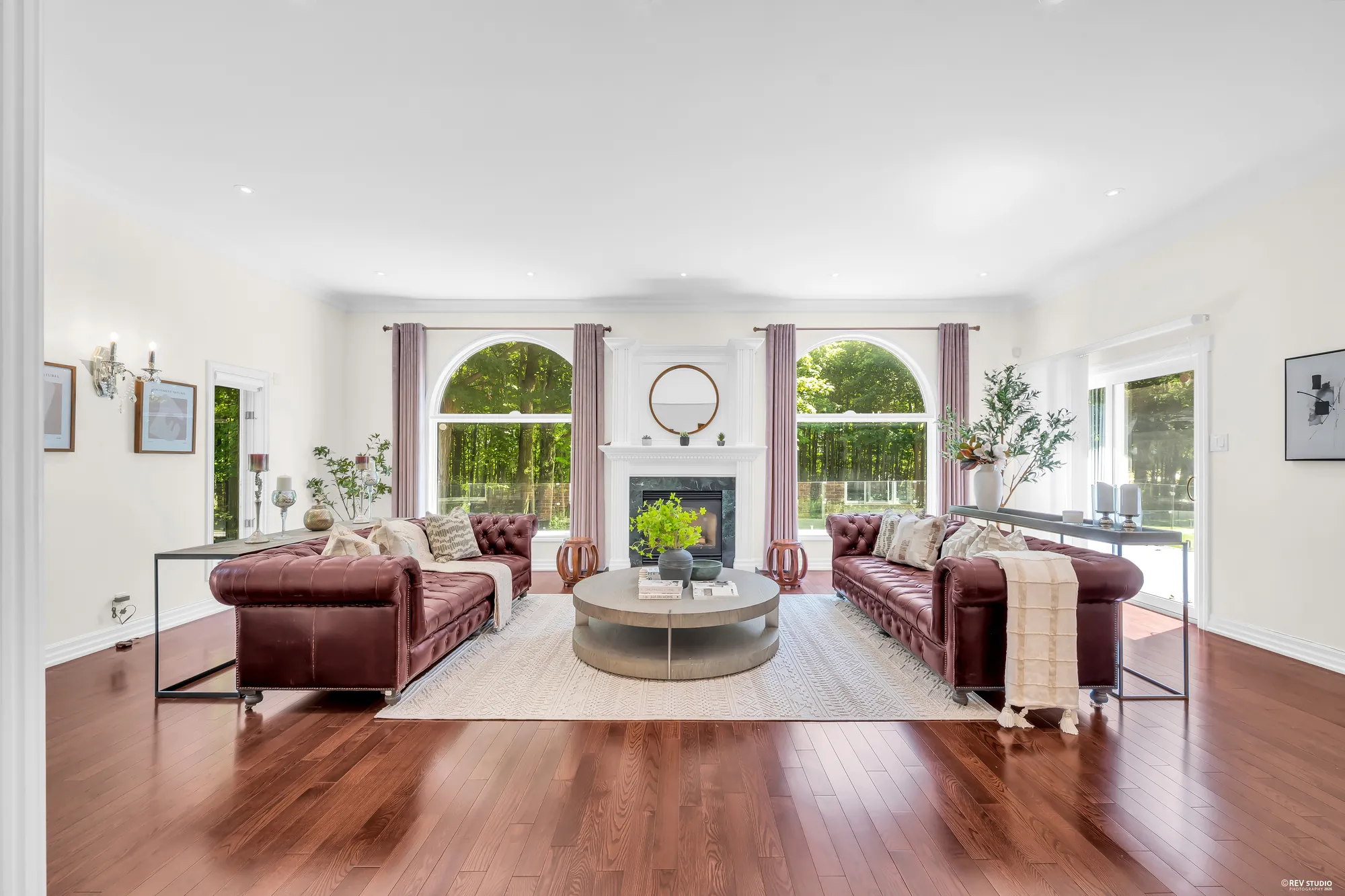 The family room: a fireplace with a white mantel and a round mirror between two tall arched windows, burgundy leather sofas facing each other across a round coffee table, a rug, two console tables