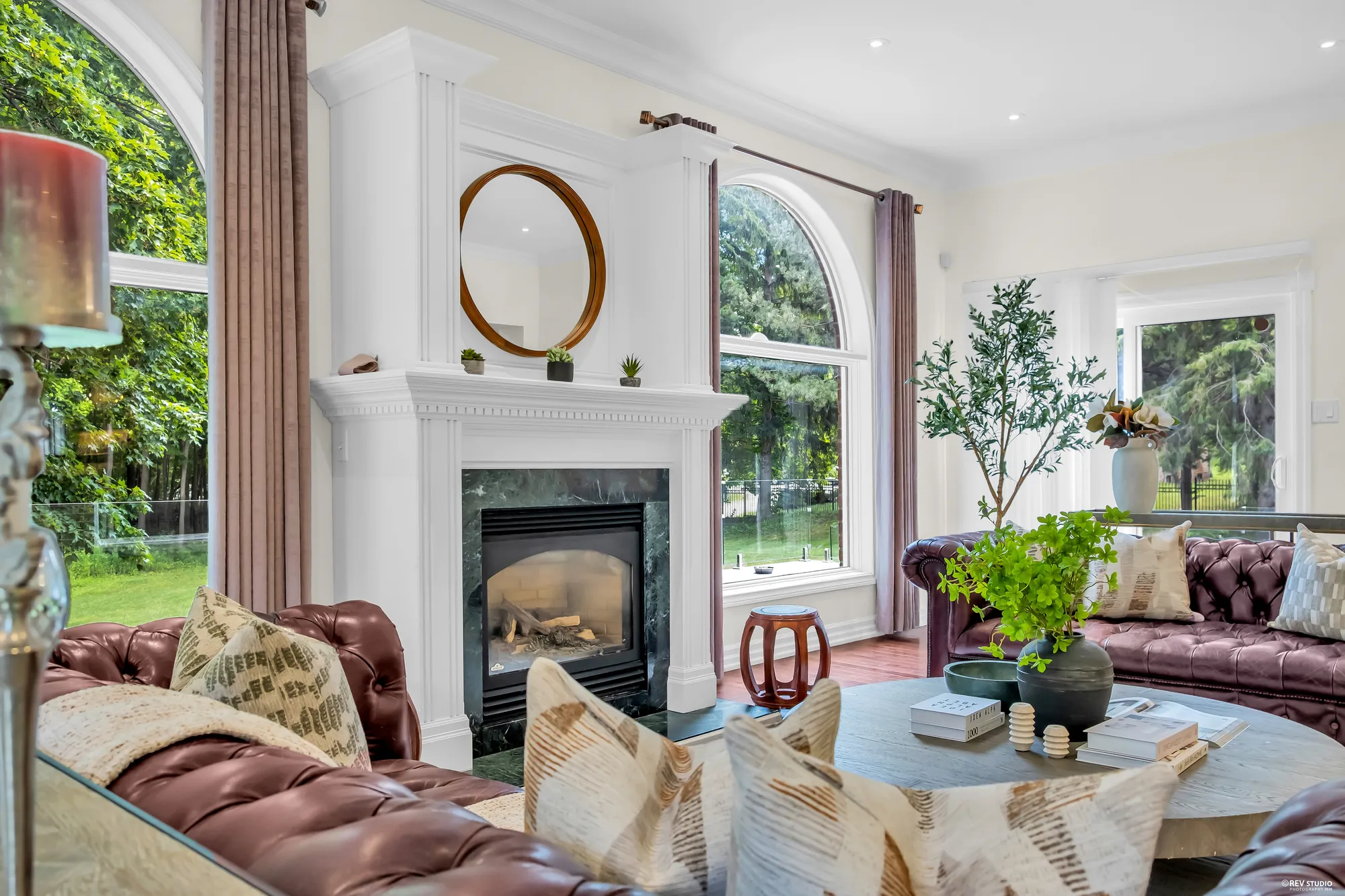 The fireplace in its dark stone surround under a white mantel, a round mirror, the arched windows, a leather sofa with cushions