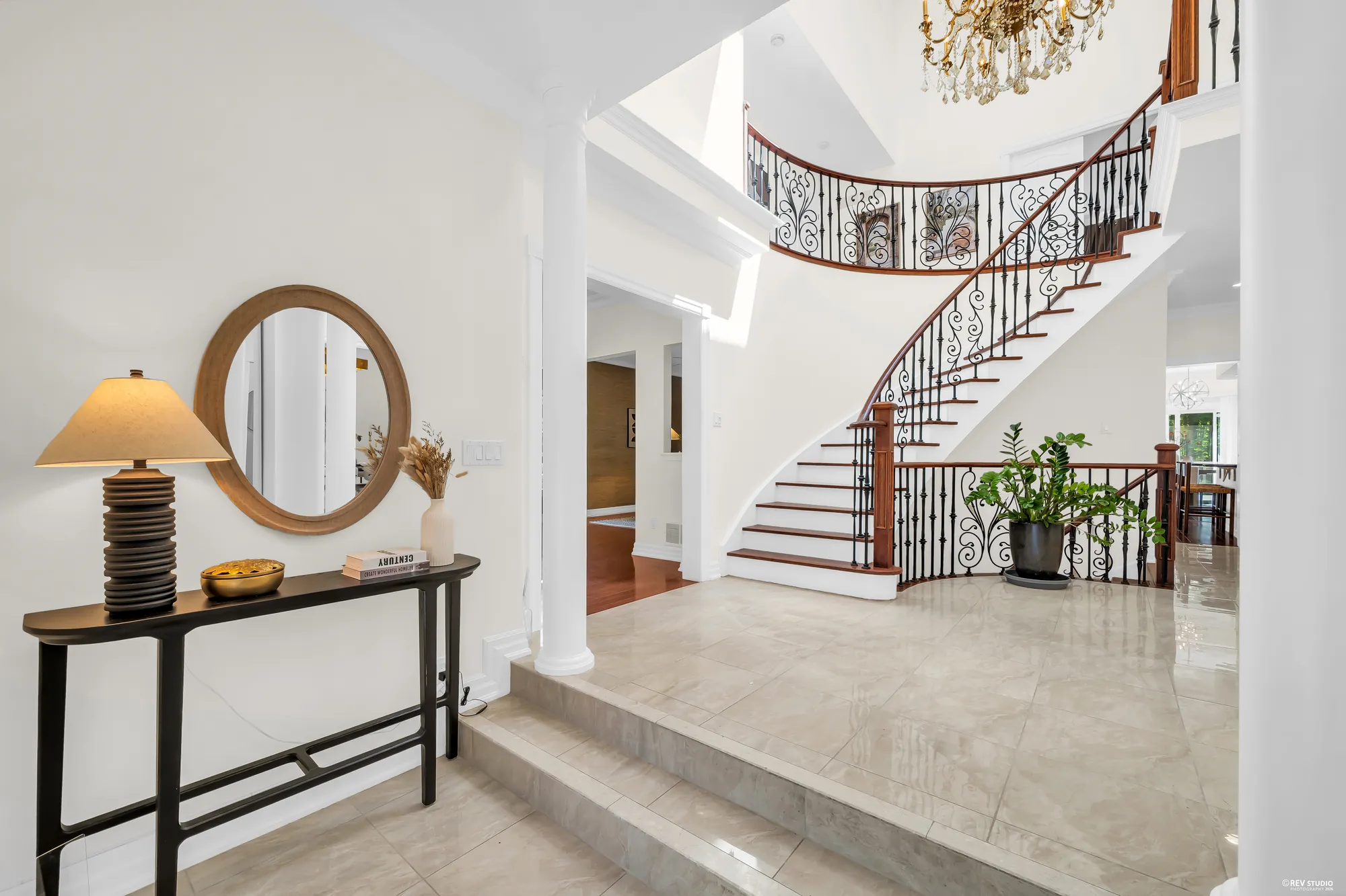 A console table with a lamp and a round mirror, a white column and a step, the curved stair beyond