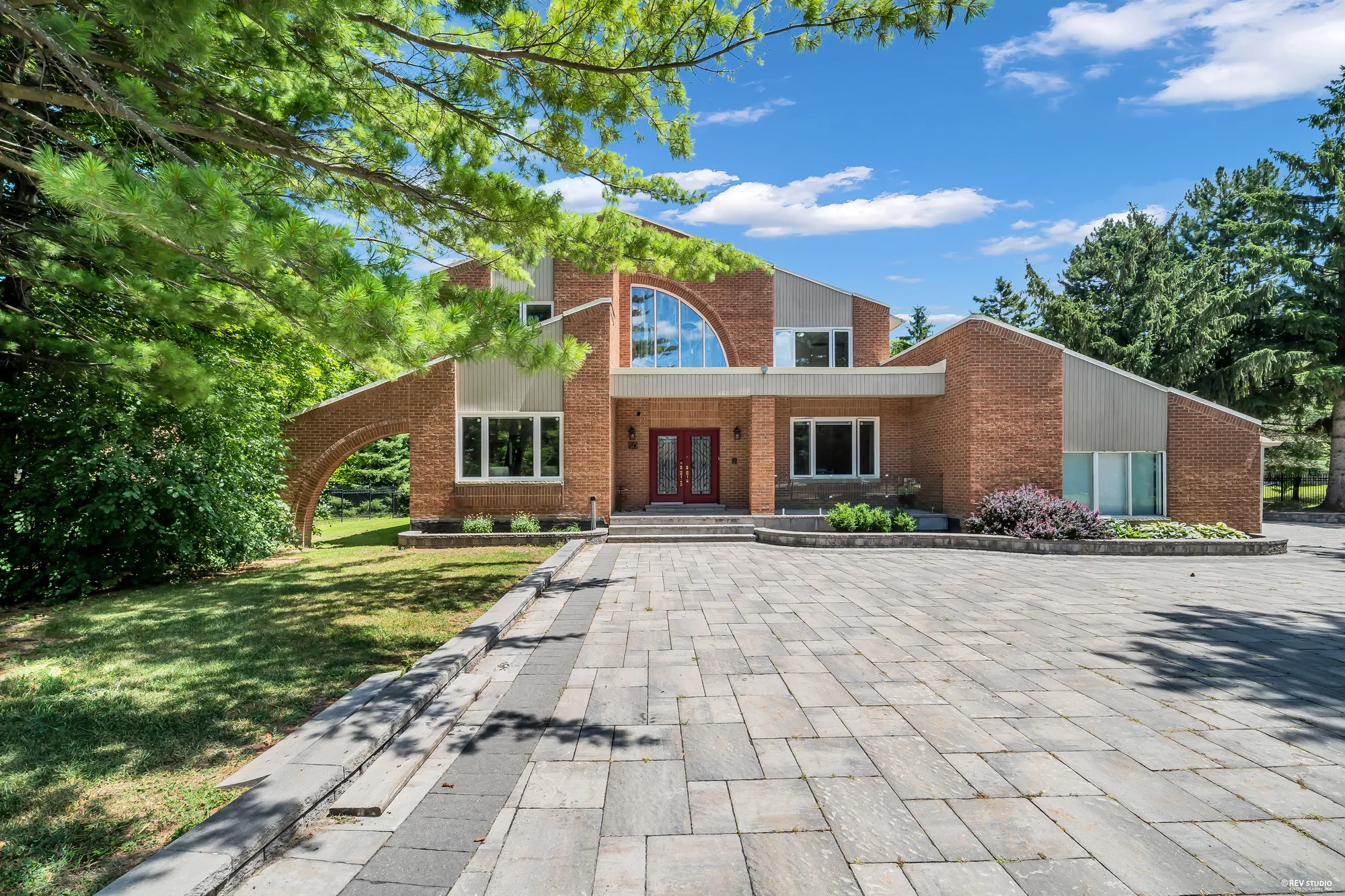 The front of the house across the paved drive: two storeys of red brick with grey siding panels, a quarter-round arched window over the entrance, a red double door under a covered entry, a brick arch reaching out from the wing at the left, a pine at the upper left