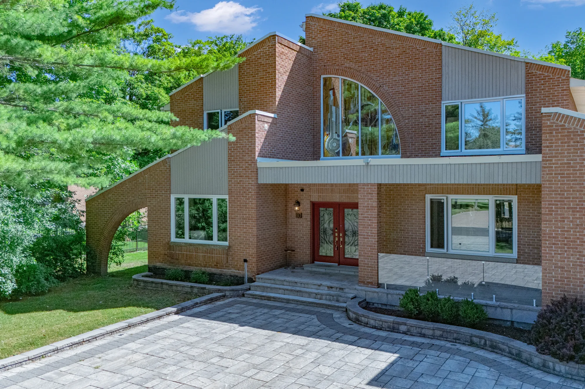 The front from a little above the drive: the quarter-round arched window between brick gables, grey siding panels, the covered entry and the red front door, the brick arch at the left