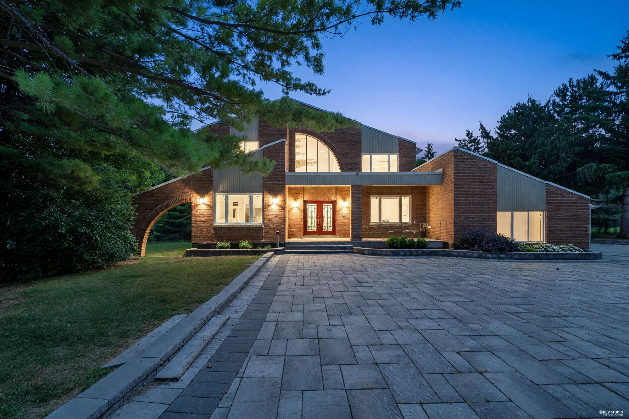 The front of 50 Boynton Circle at dusk across the paved drive: two storeys of red brick with grey siding panels, the quarter-round arched window and the other windows lit, lights on either side of the red double door, a brick arch reaching out from the wing at the left