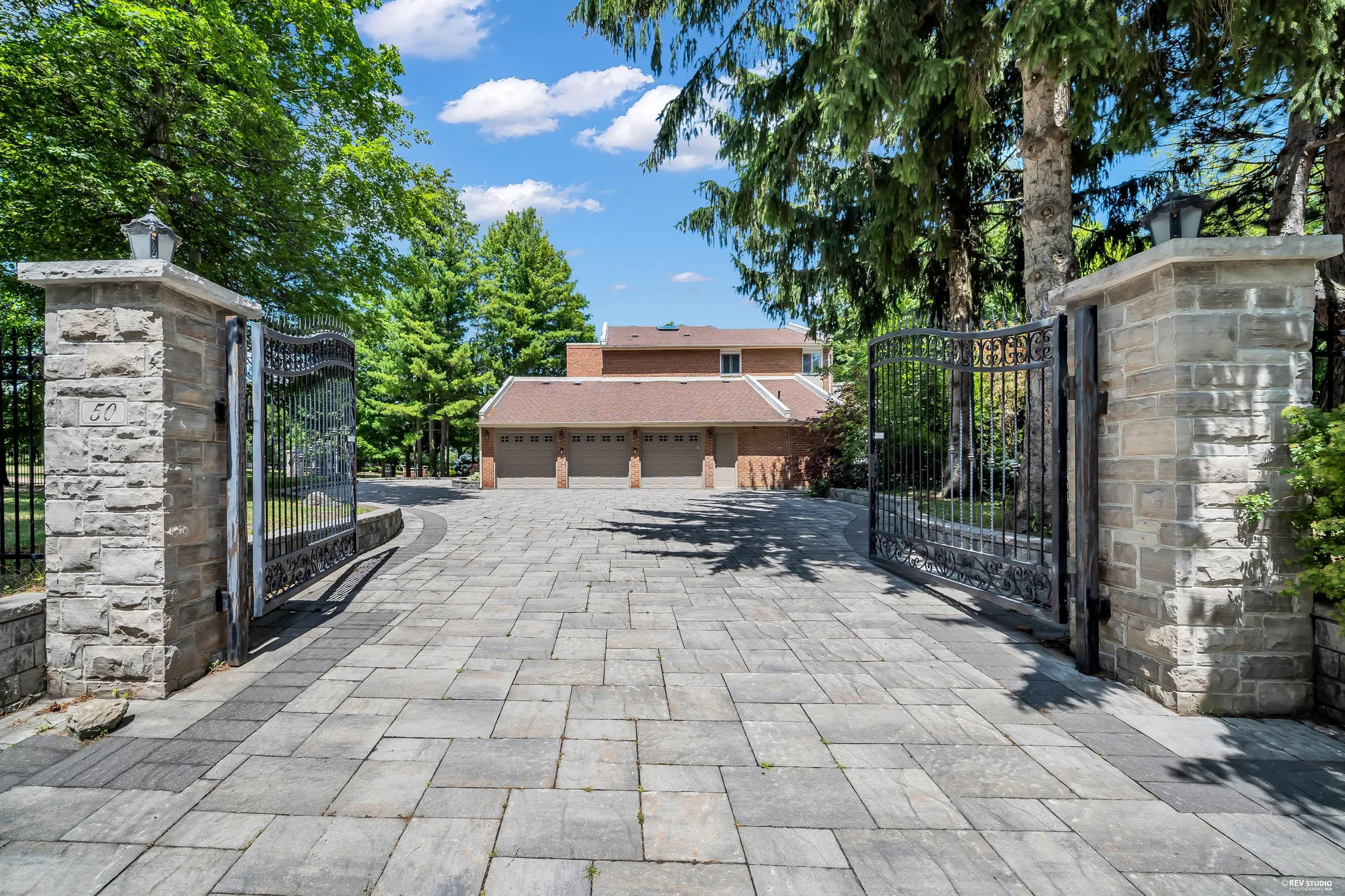 Inside the gates: stone pillars and open iron gates, a paved drive to the attached garage with three doors, tall pines behind