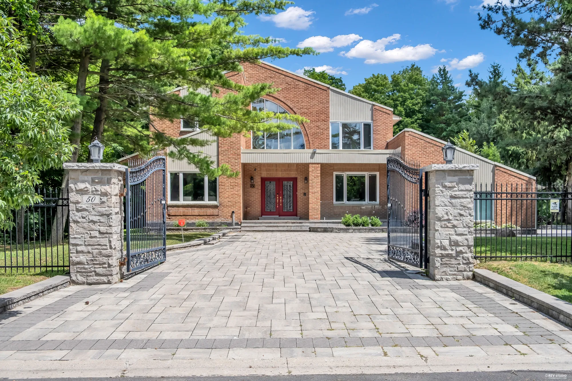 The house through the open iron gates: stone pillars with lanterns, the house number 50 on the left pillar, a paved drive to the front door, an iron fence along the lawn