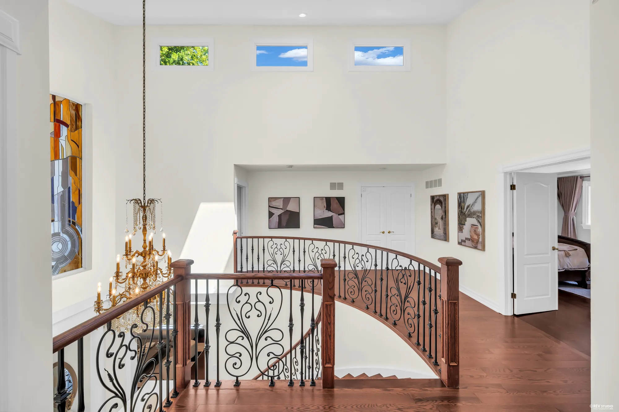 The landing at the top of the curved stair: an iron railing, a crystal chandelier, small windows high in the wall, framed prints, white double doors