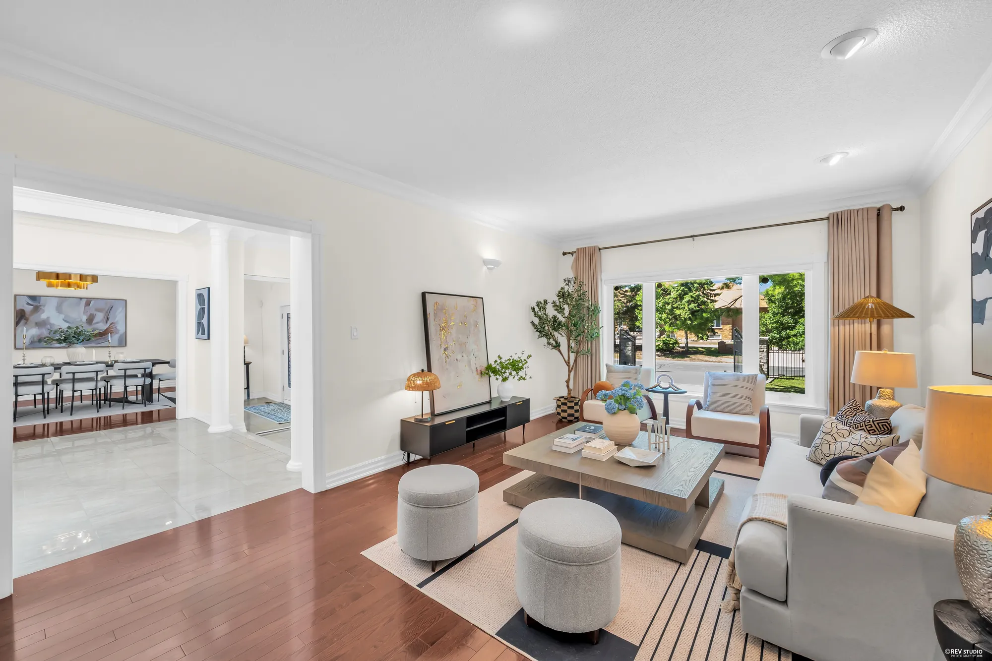 The living room: a light grey sofa and armchairs, a square coffee table and round upholstered ottomans on a rug, a wide window onto the front lawn, the front dining room across the foyer at the left