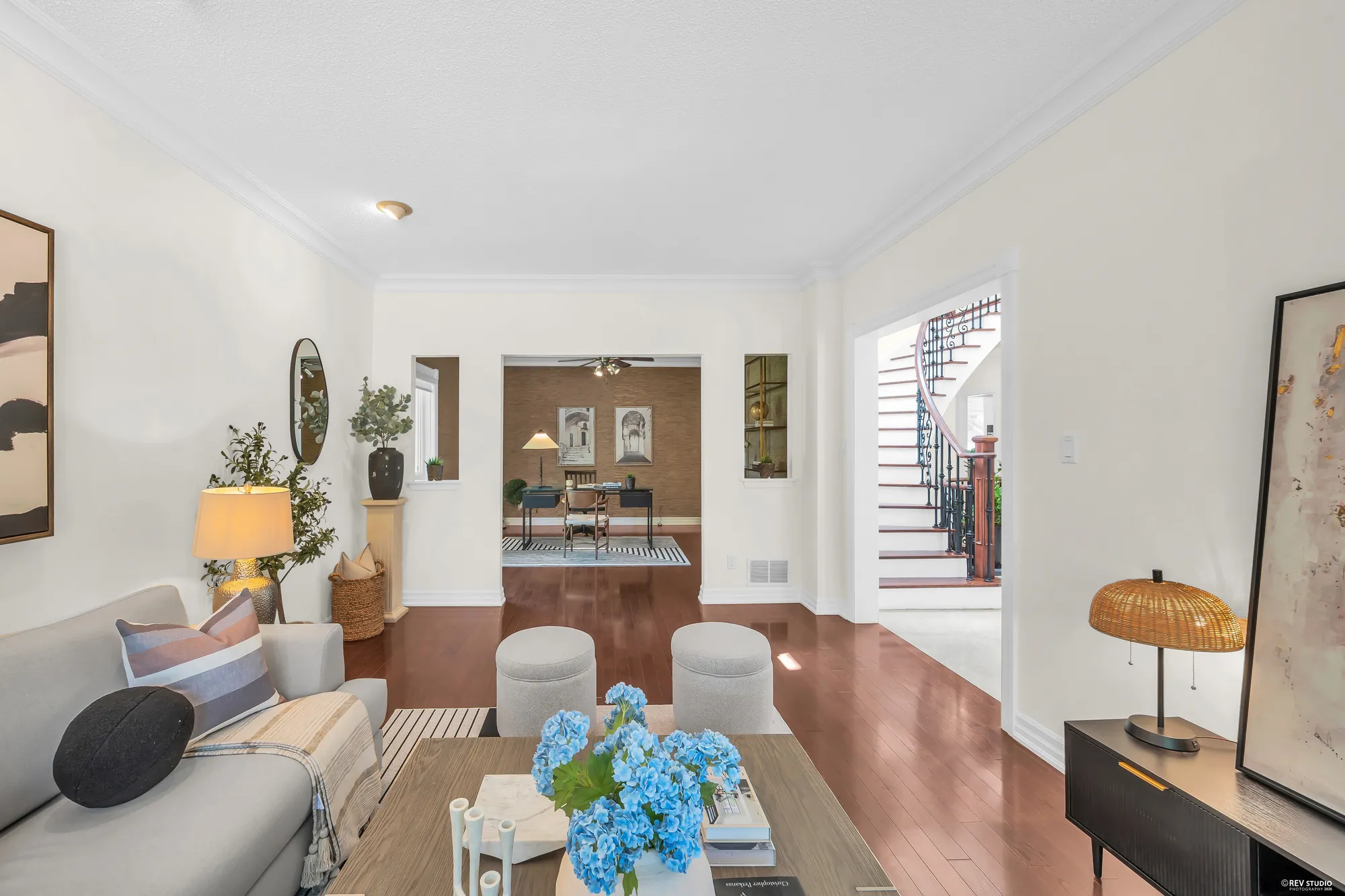 From the living room toward the office: a grey sofa, a coffee table with blue hydrangeas, the office and its desk through the doorway, the stair at the right