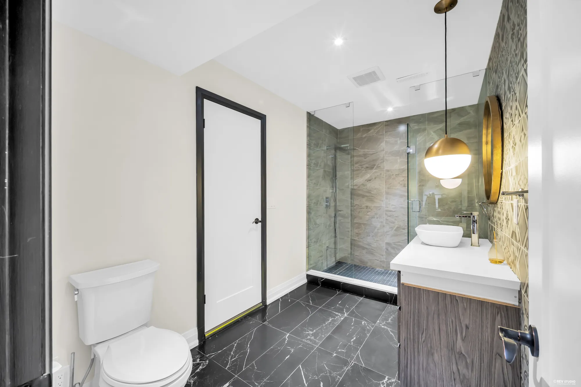 The lower bathroom: a glass shower in large grey tile, a white vessel sink on a wood vanity, a toilet, a black marble-look floor