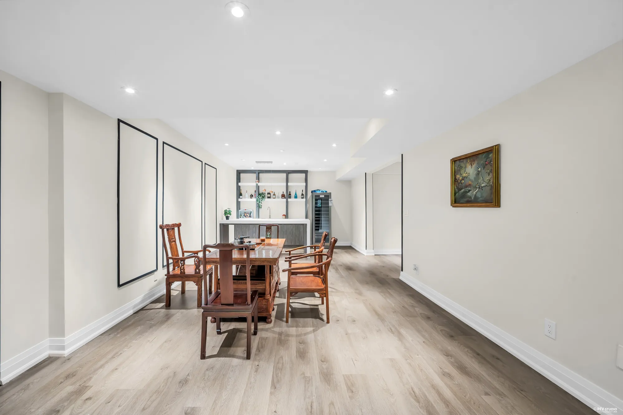 The lower dining area: a wood table with carved chairs, a painting, the wet bar at the far end with open shelves and a wine fridge