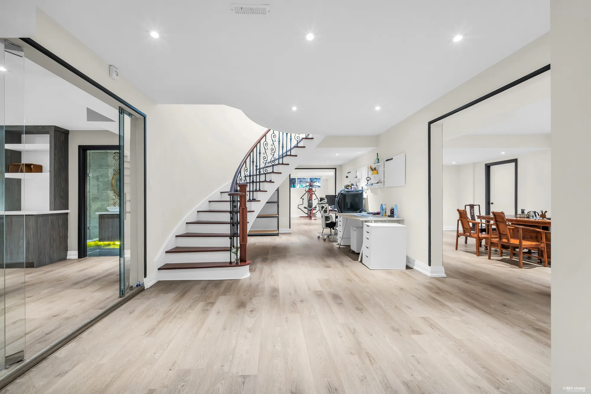 The lower hall: the curved stair down from the main floor, desks under it, the dining area through a wide opening at the right, a glass door at the left