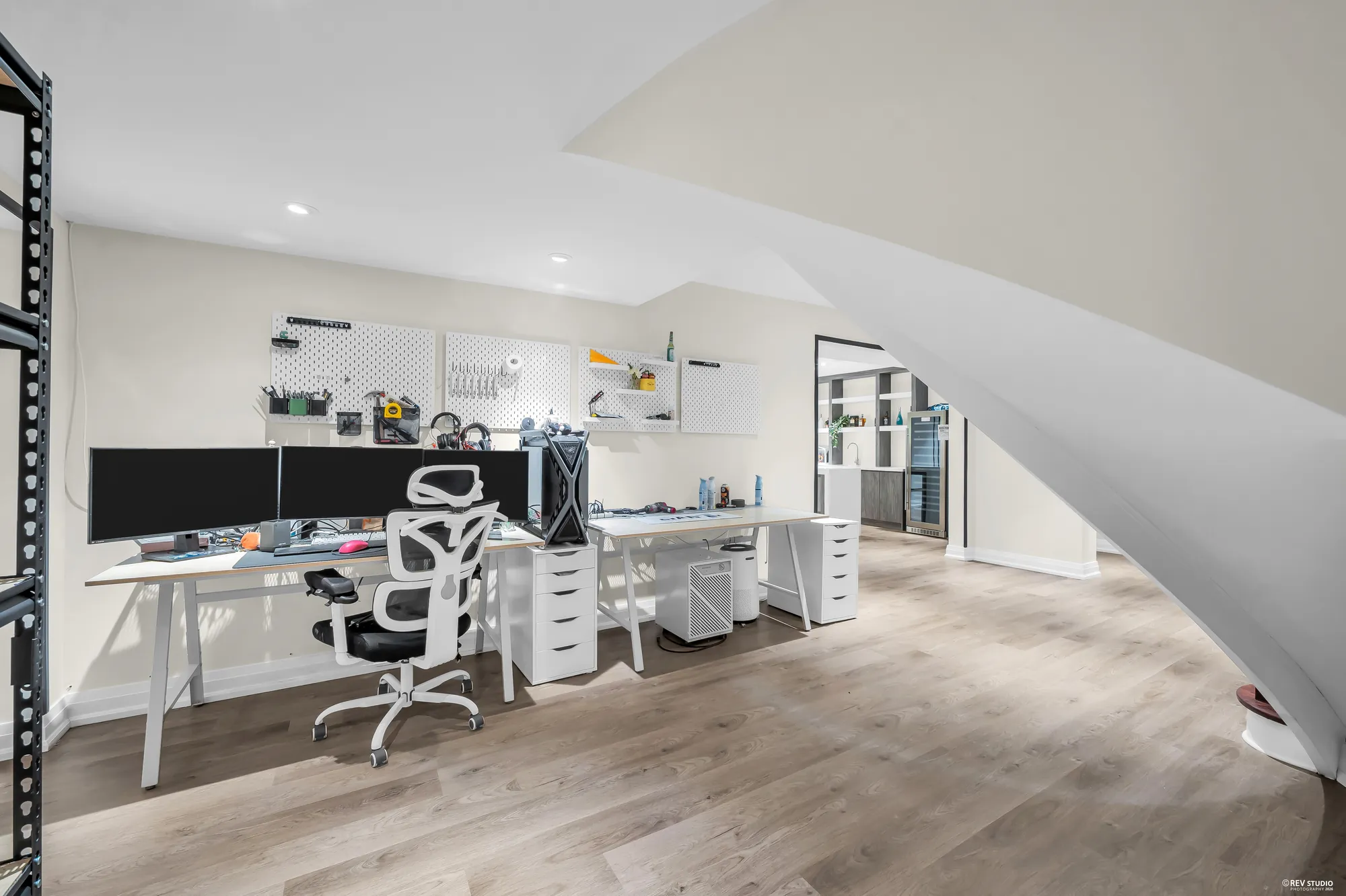 Desks with monitors and an office chair under the curve of the stair, a pegboard on the wall