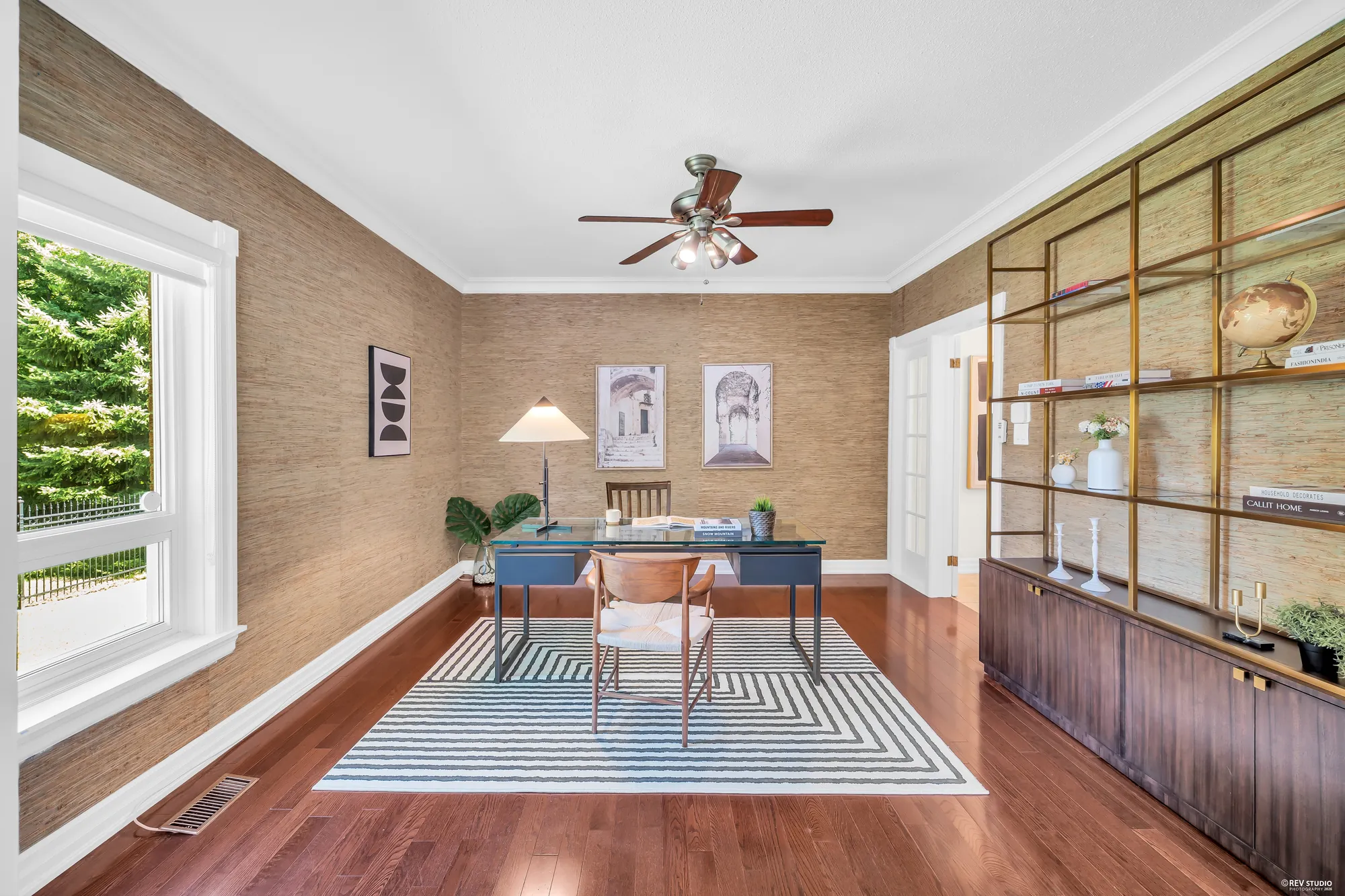 The office: grasscloth walls, a ceiling fan, a desk and chair on a striped rug, a wall of shelving in brass and dark wood, a window onto the garden