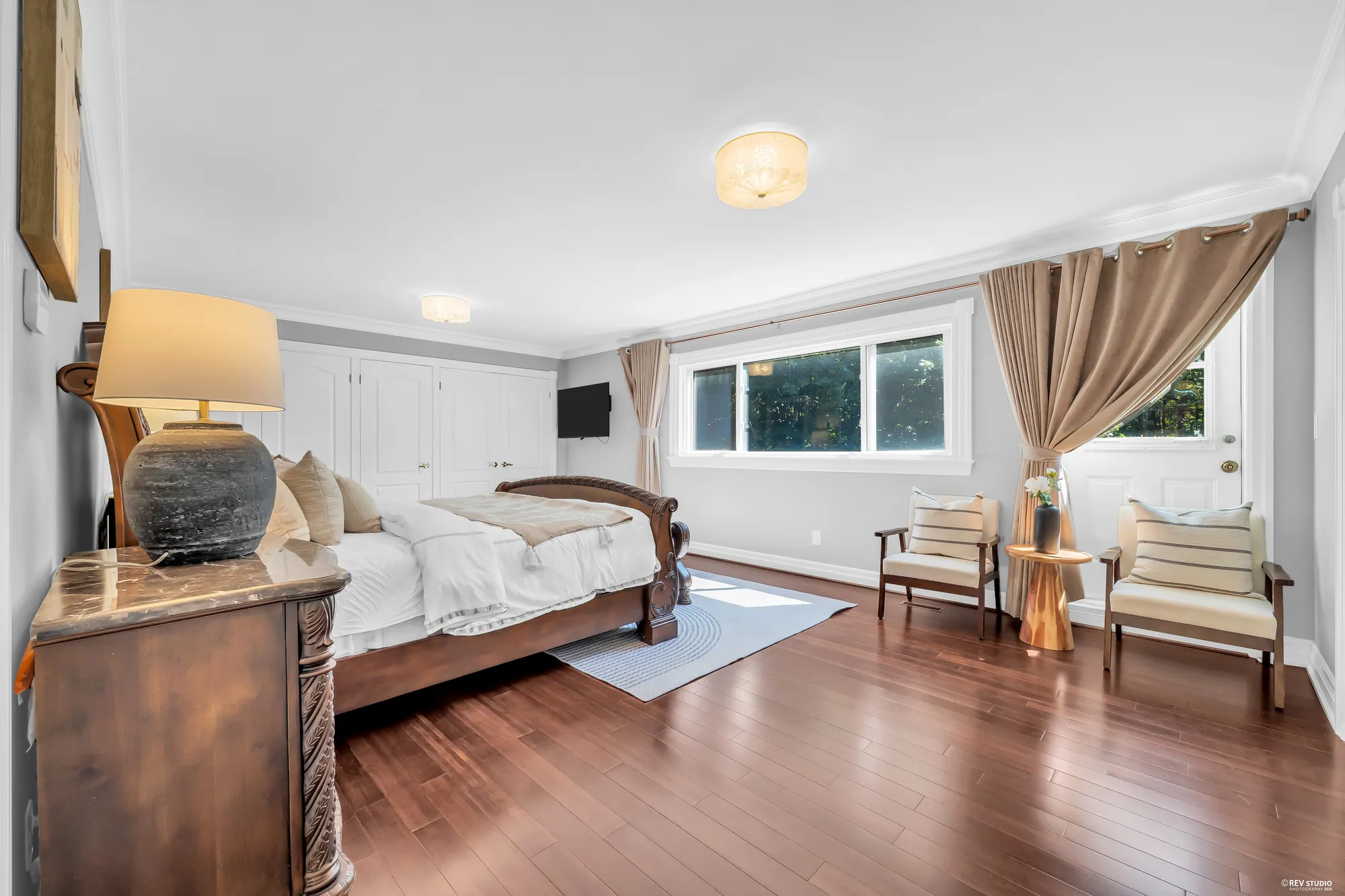 The primary bedroom: a carved wood bed, a dresser with a lamp, a wide window with tan curtains, two armchairs, a door to the balcony