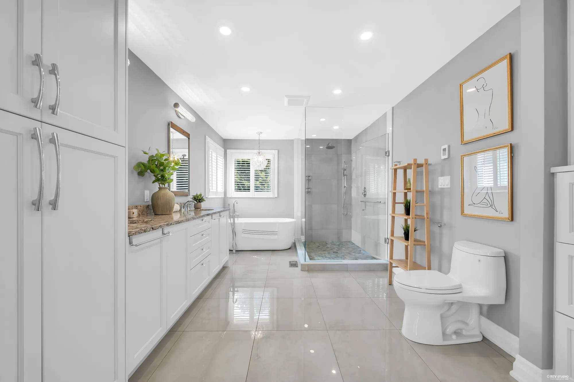 The primary bathroom: a long white vanity with a granite top, a freestanding tub under a window, a glass shower, a toilet, framed prints