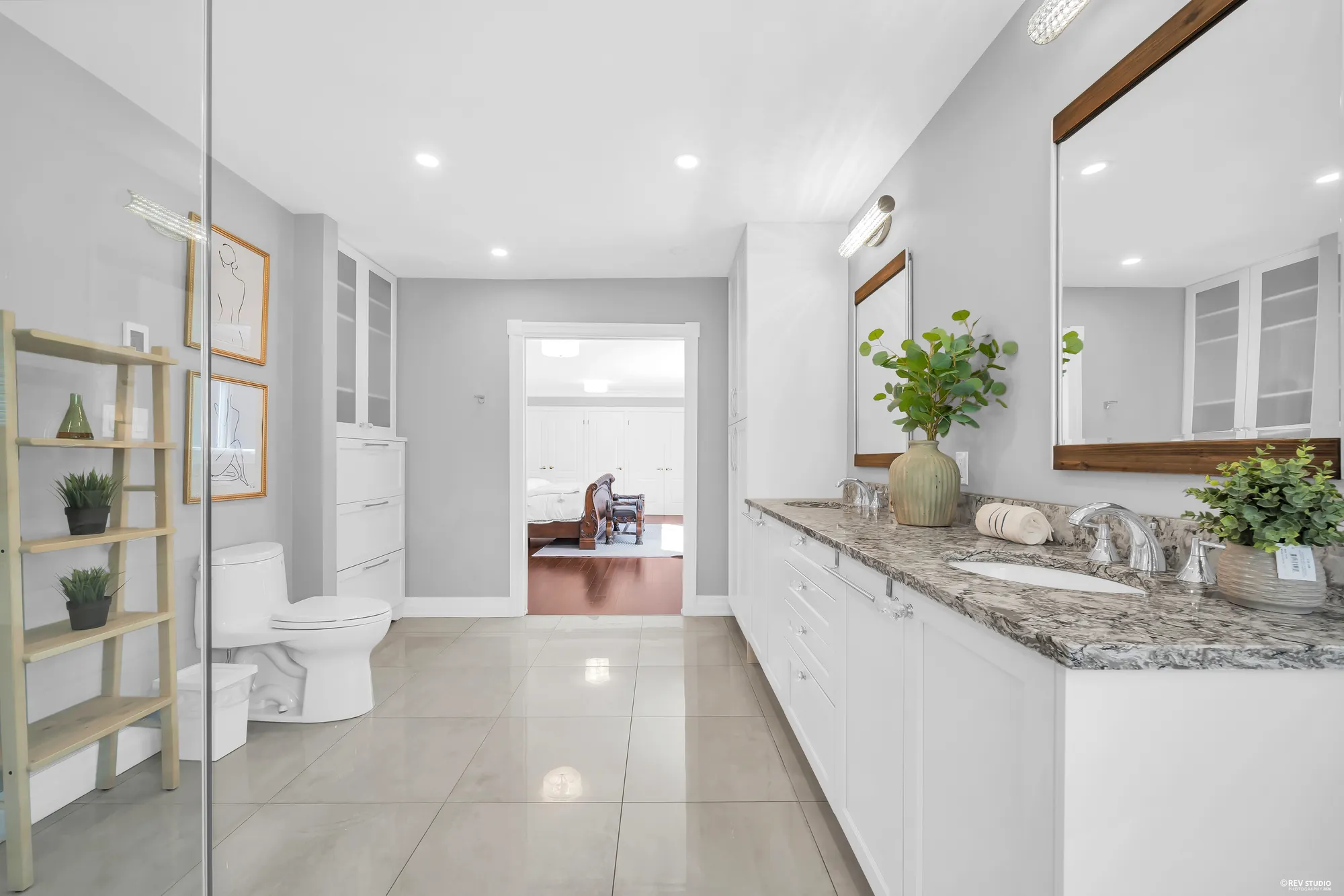 The double vanity with its mirrors and white cabinets, the glass shower at the left, the bedroom through the door