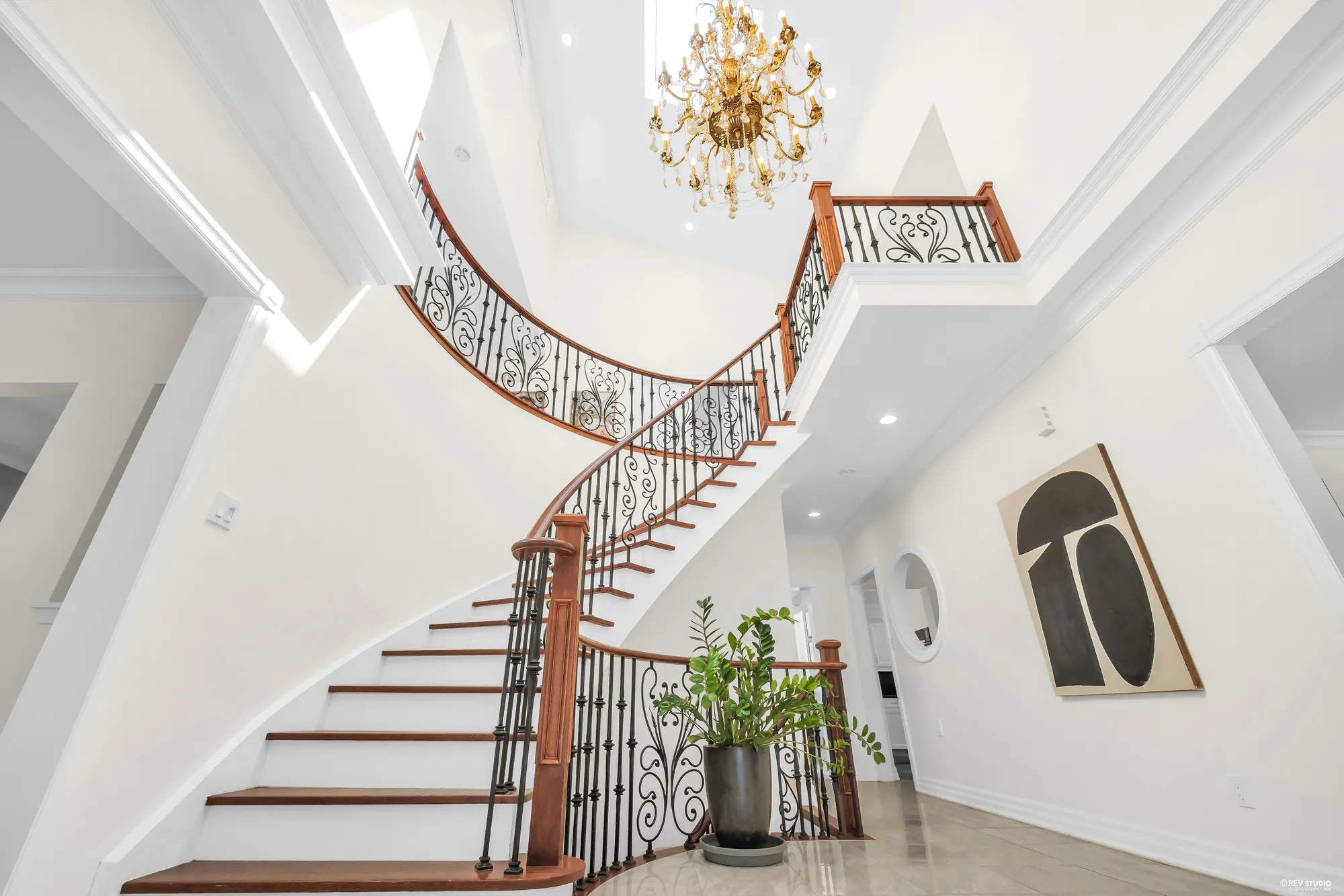 The curved stair rising from the foyer: white risers, wood treads and handrail, iron balusters, a crystal chandelier hanging in the two-storey space, a large black and cream print